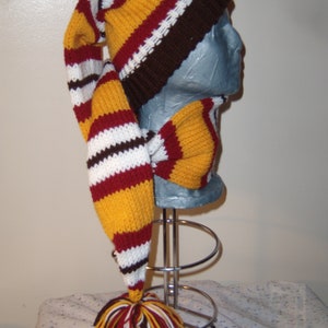 May include: A long, knitted stocking cap featuring horizontal stripes in mustard yellow, maroon, white, and dark brown. The cap has a ribbed dark brown cuff and a large, multi-colored yarn pom-pom attached to the pointed end of the long tail.