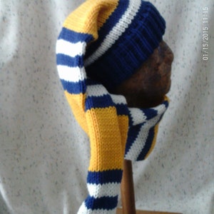 May include: A knitted winter hat with a long, pointed tail and a pom-pom. The hat features a color scheme of yellow, blue, and white stripes. The hat is displayed on a wooden head form.