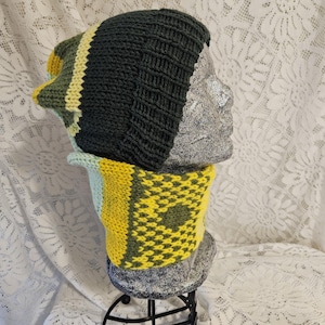 May include: A knitted beanie and matching cowl set featuring a dark charcoal gray ribbed beanie and a cowl with a geometric pattern in yellow, olive green, and light blue. The set is displayed on a textured gray mannequin head and neck form.