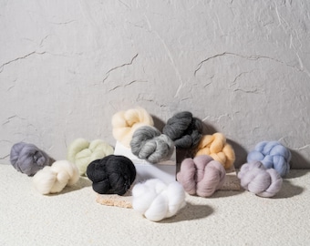 May include: A collection of colorful wool roving in various shades, including cream, gray, black, and pastel hues. The wool is arranged in fluffy, cloud-like bundles, perfect for fiber art projects. The background is a textured, light gray wall.