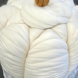 May include: A large, white, fluffy pumpkin-shaped creation made of thick wool roving. A small, orange pumpkin with a green stem sits on top. The wool is tightly packed and appears soft.