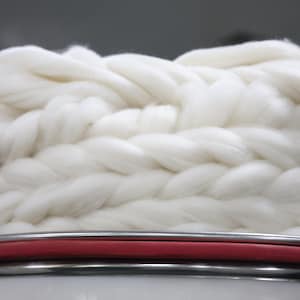 May include: Close-up of a thick, white, braided yarn. The yarn has a soft, fluffy texture. Below the yarn is a red and silver metal bar. The background is a blurred grey.
