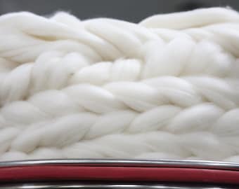 May include: Close-up of a thick, white, braided yarn. The yarn is a soft, fluffy texture. Below the yarn is a red and silver metal bar. The background is a blurred gray.