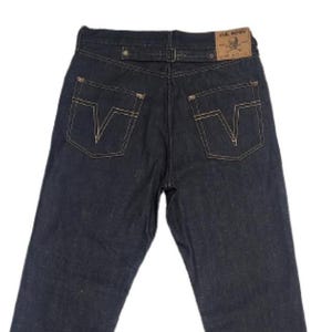Puede incluir: Vaqueros de mezclilla azul oscuro con pernera recta y puños doblados. Los bolsillos traseros presentan un diseño en forma de V con costuras en contraste. Un parche de cuero es visible en la cintura.