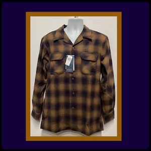 Puede incluir: Una camisa de manga larga de cuadros marrones y negros con cuello y cierre de botones. La camisa tiene dos bolsillos en el pecho y una etiqueta que dice "Made in USA".