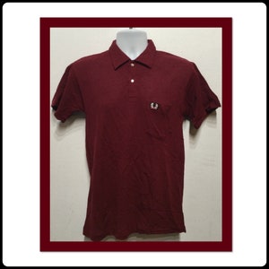 May include: A maroon polo shirt with a small embroidered logo on the left chest. The logo is a white laurel wreath with a red circle around it.