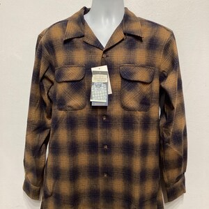 Puede incluir: Una camisa de manga larga de cuadros marrones y negros con cuello abotonado y dos bolsillos en el pecho. La camisa tiene un aspecto descolorido y est&aacute; colgada de una percha. La etiqueta de la marca dice "Pendleton".
