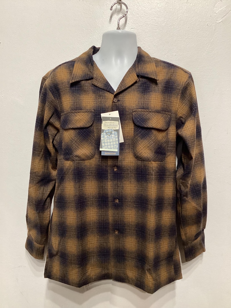 Puede incluir: Una camisa de manga larga de cuadros marrones y negros con cuello abotonado y dos bolsillos en el pecho. La camisa tiene un aspecto descolorido y est&aacute; colgada de una percha. La etiqueta de la marca dice "Pendleton".