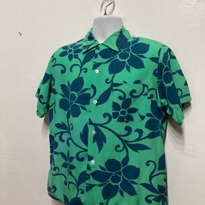 May include: Green short-sleeve shirt with a large floral pattern in dark blue. The shirt has a collared neckline, a chest pocket, and white buttons. The floral design is repeated across the fabric.