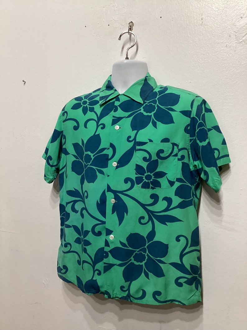 May include: Green short-sleeve shirt with a large floral pattern in dark blue. The shirt has a collared neckline, a chest pocket, and white buttons. The floral design is repeated across the fabric.