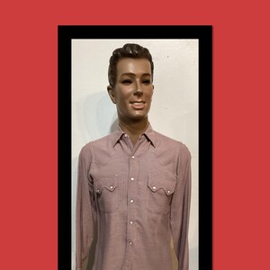 May include: A long-sleeved button-down shirt in a muted mauve and white micro-check pattern, featuring a pointed collar, two chest pockets with pointed flaps, and white snap buttons, displayed on a male mannequin with dark brown hair.