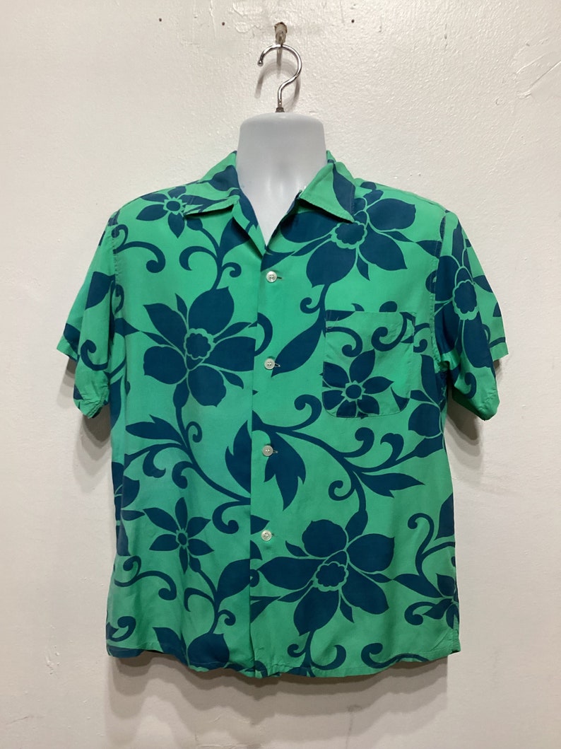 May include: A short-sleeved button-down shirt in a vibrant green hue, adorned with a bold floral pattern in a contrasting dark teal. The shirt features a classic collar, a chest pocket, and white buttons.