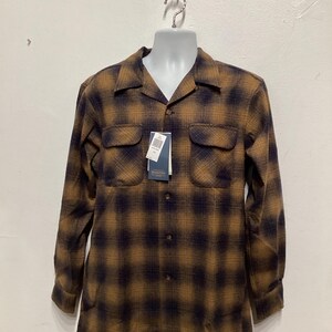 Puede incluir: Una camisa de manga larga con botones y un estampado de cuadros marrones y negros. La camisa tiene cuello y dos bolsillos en el pecho.