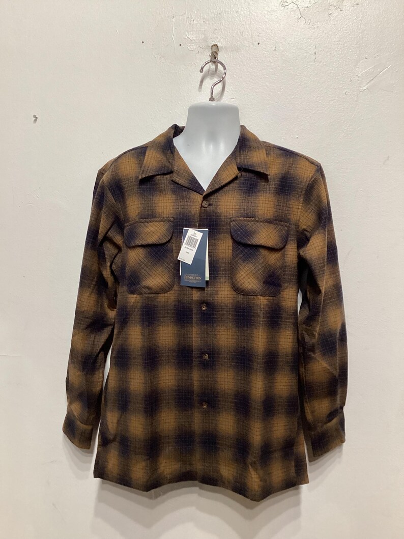Puede incluir: Una camisa de manga larga con botones y un estampado de cuadros marrones y negros. La camisa tiene cuello y dos bolsillos en el pecho.