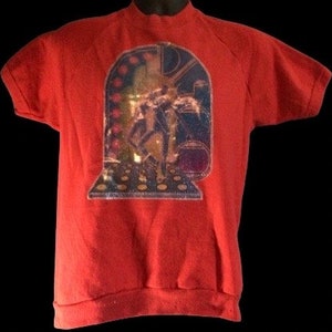 May include: Red short-sleeved sweatshirt with a colourful graphic of a person in a doorway. The graphic has a glittery finish.