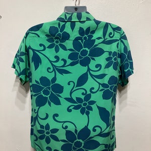 May include: Green short-sleeved shirt with a teal floral pattern. The shirt is hanging on a hanger. The floral design features large flowers and swirling vines, creating a tropical aesthetic.