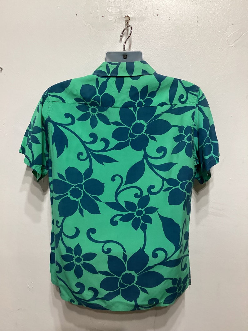 May include: Green short-sleeved shirt with a teal floral pattern. The shirt is hanging on a hanger. The floral design features large flowers and swirling vines, creating a tropical aesthetic.