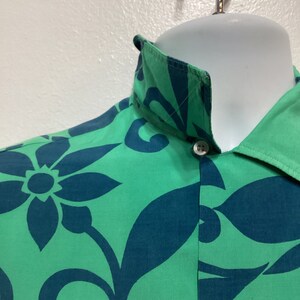 May include: Close-up of a green button-down shirt with a teal floral pattern. The shirt is displayed on a white mannequin head and features a classic collar and white buttons. The fabric has a smooth texture.