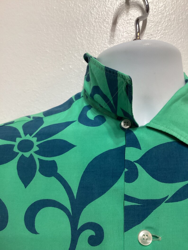 May include: Close-up of a green button-down shirt with a teal floral pattern. The shirt is displayed on a white mannequin head and features a classic collar and white buttons. The fabric has a smooth texture.