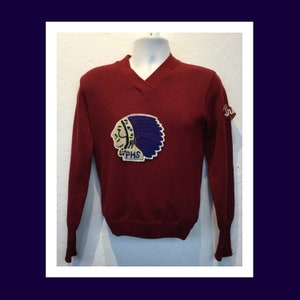 Puede incluir: Un suéter vintage de color granate con un gran parche bordado de la cabeza de un indio americano con un tocado. El parche también tiene las letras "PHS".