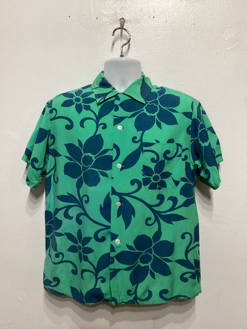 May include: A short-sleeved button-down shirt in a vibrant green hue, adorned with a bold floral pattern in a contrasting deep teal. The shirt features a classic collar, a chest pocket, and white buttons.