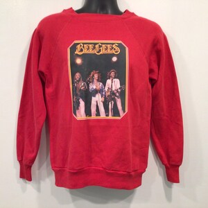 Könnte beinhalten: Rotes Sweatshirt mit einer Grafik der Bee Gees. Die Grafik ist rechteckig mit einem goldenen Rand und dem Namen der Band oben. Das Sweatshirt hat lange Ärmel und einen Rundhalsausschnitt.