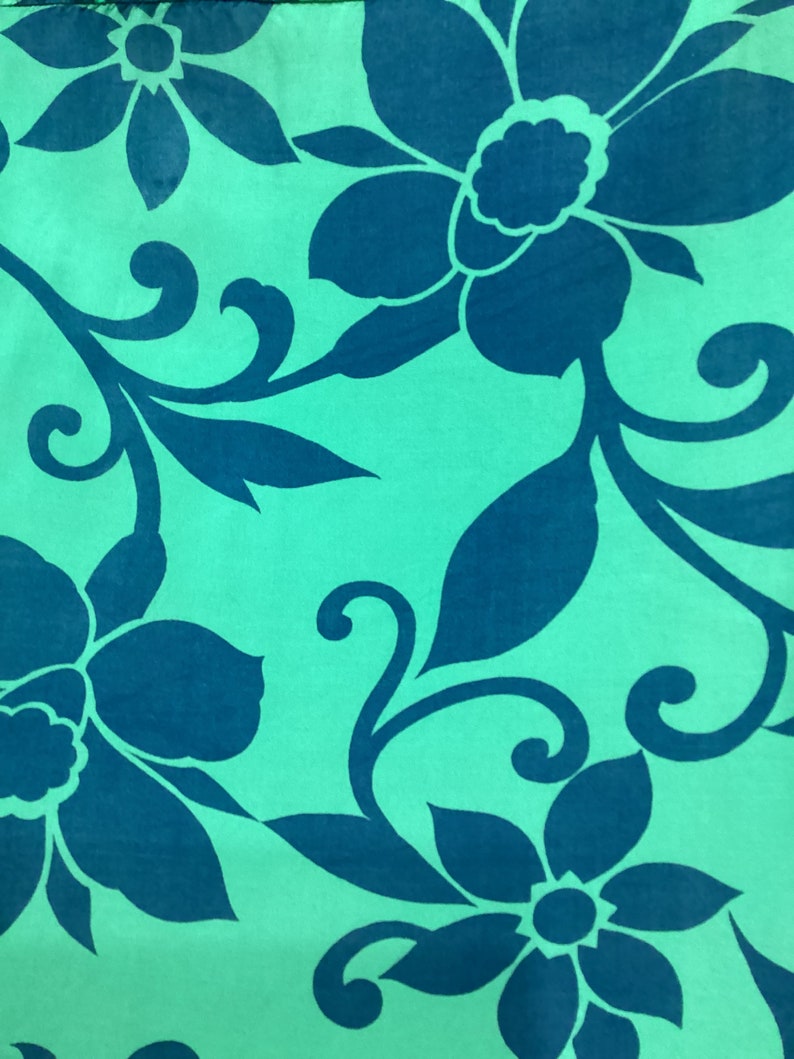 May include: A close-up of a fabric with a repeating floral pattern. Large, stylized flowers and leaves in a deep teal color are set against a light teal background. The design features curved stems and swirling accents.