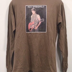 May include: A brown long-sleeve t-shirt with a graphic of Rick Springfield holding a guitar. The graphic is in a black frame with the text "Rick Springfield" above it.
