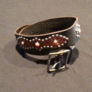 May include: A dark brown leather belt with silver-colored studs and decorative accents. The belt features a rectangular silver buckle and is adorned with small, round studs and oval-shaped, orange-colored stones.
