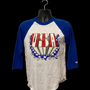 May include: A white and blue baseball t-shirt with a red, white, and blue graphic of the word "USA" with stars and stripes. The graphic is set against a ripped background.