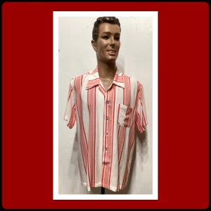 May include: A short-sleeved, button-down shirt with vertical red stripes on a white background. The shirt has a single chest pocket and a classic collar. The shirt is displayed on a mannequin.