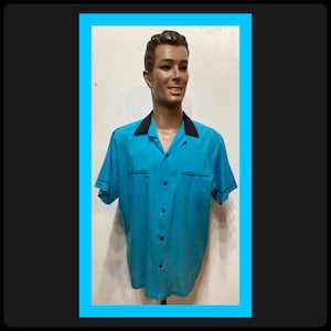 Può includere: Una camicia turchese a maniche corte con colletto e finiture nere. La camicia ha due tasche sul petto e bottoni neri. La camicia è esposta su un manichino.