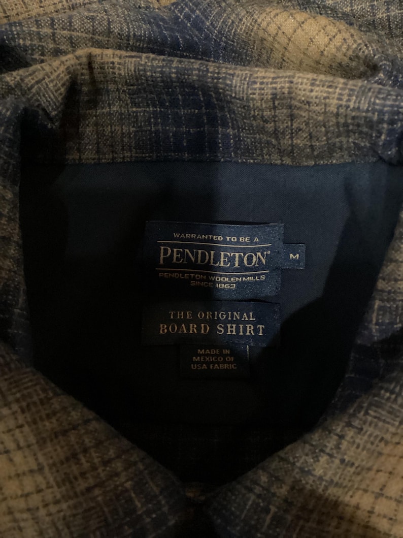 Puede incluir: Una camisa Pendleton de cuadros marrones y azules con una etiqueta que dice "WARRANTED TO BE A PENDLETON PENDLETON WOOLEN MILLS SINCE 1963 THE ORIGINAL BOARD SHIRT MADE IN MEXICO OF USA FABRIC" y una etiqueta de talla que dice "M".
