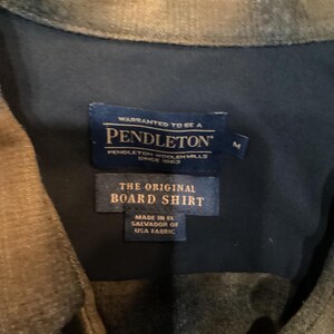 以下が含まれることがあります： ペンドルトンボードシャツのラベルのクローズアップ。ラベルには「Warranted to be a Pendleton, Pendleton Woolen Mills Since 1863」と書かれています。その下に、「The Original Board Shirt, Made in El Salvador of USA Fabric」と書かれた2番目のラベルがあります。