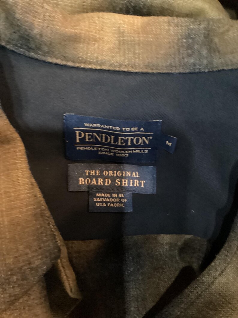 以下が含まれることがあります： ペンドルトンボードシャツのラベルのクローズアップ。ラベルには「Warranted to be a Pendleton, Pendleton Woolen Mills Since 1863」と書かれています。その下に、「The Original Board Shirt, Made in El Salvador of USA Fabric」と書かれた2番目のラベルがあります。