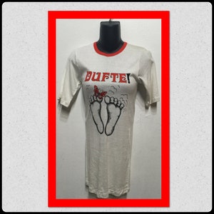 May include: White short-sleeved t-shirt with a red and black ringer neck. The shirt features a graphic of two feet with a butterfly on top and the word "DUFTE!" printed in red.