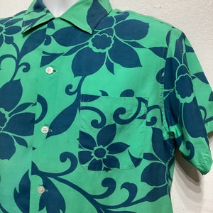 May include: Green short-sleeve shirt with a large floral pattern in teal. The shirt has a button-down front, a chest pocket, and a classic collar. The fabric appears to be lightweight.