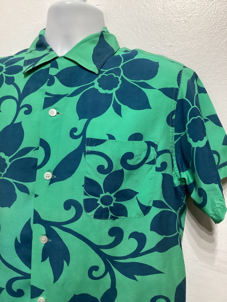 May include: Green short-sleeve shirt with a large floral pattern in teal. The shirt has a button-down front, a chest pocket, and a classic collar. The fabric appears to be lightweight.