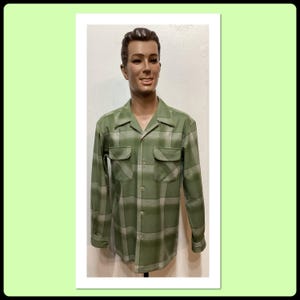 Könnte beinhalten: Ein grünes kariertes Hemd mit Knöpfen, zwei Brusttaschen und langen Ärmeln. Das Hemd wird auf einer Schaufensterpuppe präsentiert. Das Karomuster kombiniert grüne und hellgraue Quadrate. Das Hemd hat einen klassischen Kragen.
