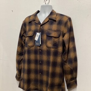Puede incluir: Una camisa de manga larga a cuadros marrones y negros con cuello y dos bolsillos en el pecho. La camisa est&aacute; colgada de un maniqu&iacute; blanco.