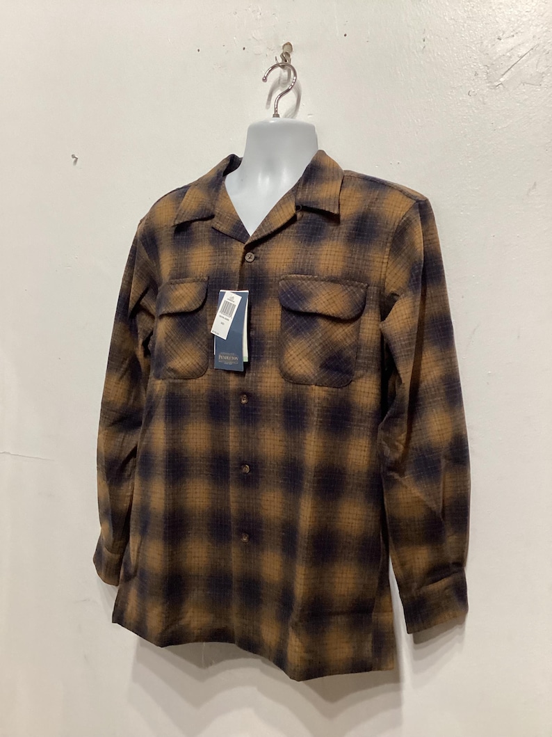 Puede incluir: Una camisa de manga larga a cuadros marrones y negros con cuello y dos bolsillos en el pecho. La camisa est&aacute; colgada de un maniqu&iacute; blanco.