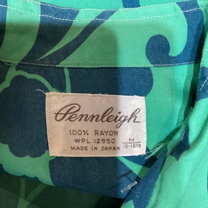 May include: Close-up of a vintage Pennleigh shirt tag. The shirt is made of 100% rayon and features a vibrant green and blue floral pattern. The tag indicates the shirt is a size M, made in Japan.