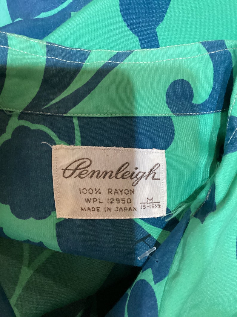 May include: Close-up of a vintage Pennleigh shirt tag. The shirt is made of 100% rayon and features a vibrant green and blue floral pattern. The tag indicates the shirt is a size M, made in Japan.