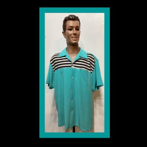 May include: A turquoise short-sleeve button-down shirt with a black and white striped design across the chest and shoulders. The shirt is displayed on a mannequin.