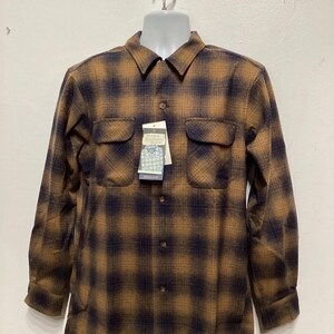 Puede incluir: Una camisa de manga larga a cuadros marrones y negros con cierre de botones en la parte delantera y dos bolsillos en el pecho. La camisa tiene un aspecto descolorido y est&aacute; colgada de una percha blanca.
