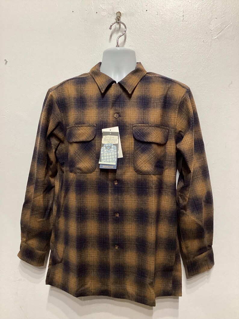 Puede incluir: Una camisa de manga larga a cuadros marrones y negros con cierre de botones en la parte delantera y dos bolsillos en el pecho. La camisa tiene un aspecto descolorido y est&aacute; colgada de una percha blanca.