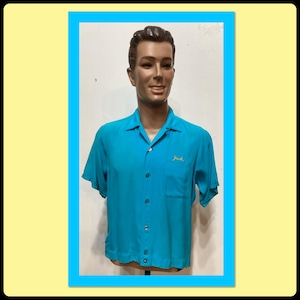 Peut inclure: Une chemise turquoise à manches courtes avec un col et une poche poitrine. Le nom "Jack" est brodé en fil d'or sur la poitrine droite. La chemise est présentée sur un mannequin.