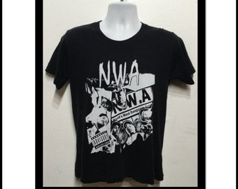May include: Black t-shirt featuring a white graphic print of the rap group N.W.A. The graphic includes the band's name, a collage of the band members' faces, and the text "World's Most Dangerous Group".