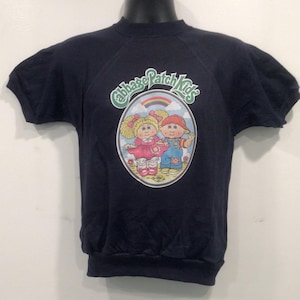 Könnte beinhalten: Ein dunkelblaues T-Shirt mit kurzen Ärmeln und einem Cabbage Patch Kids-Motiv. Das Motiv zeigt einen Regenbogen, Wolken und zwei Cabbage Patch Kids-Puppen.
