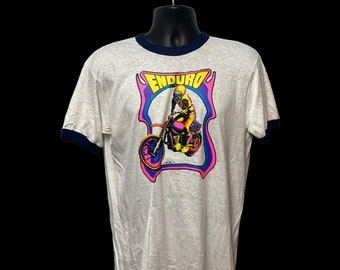 May include: White t-shirt with a blue ringer neck and a graphic of a person riding a motorcycle with the word "ENDURO" in yellow letters.